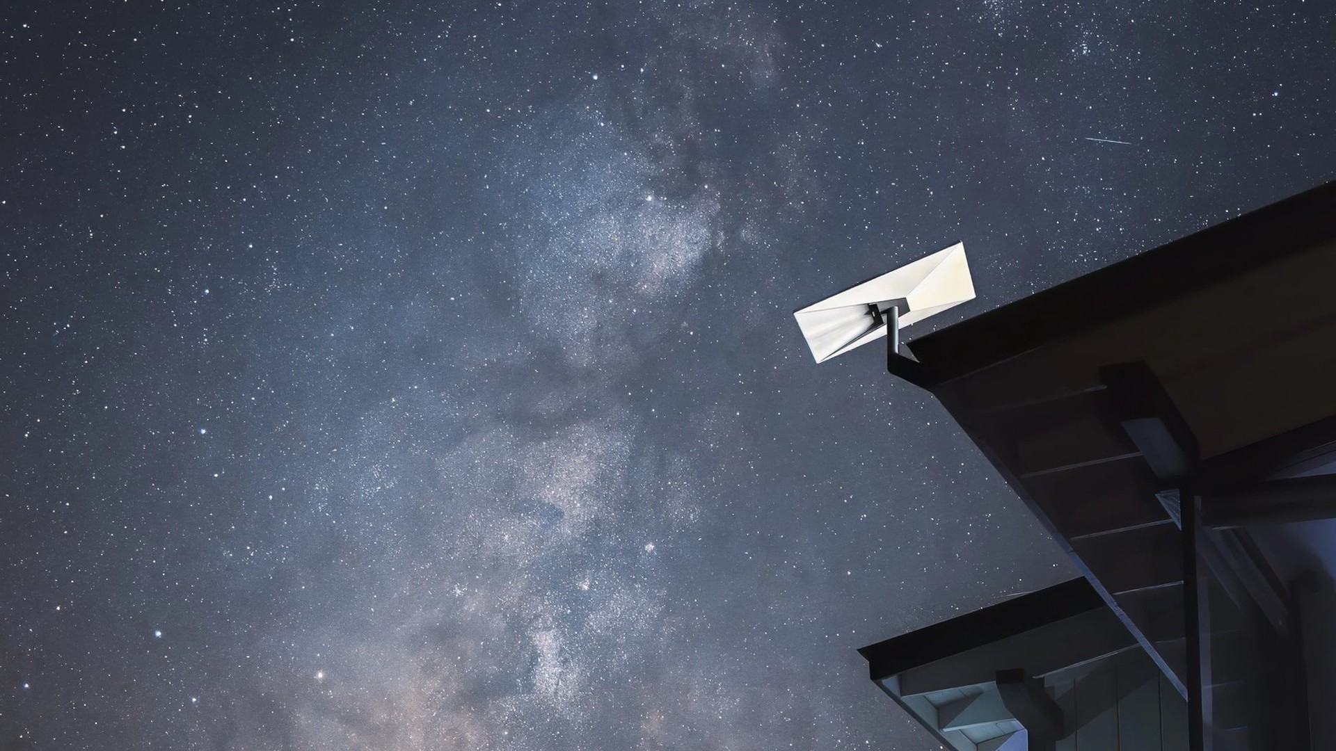 A white Starlink dish mounted on the edge of a roof under a starry night sky with the Milky Way overhead