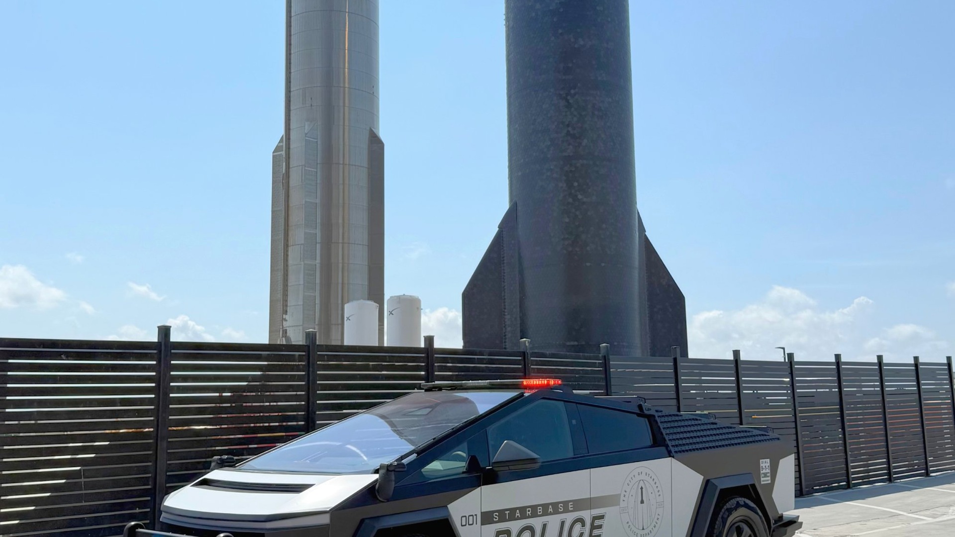Police SUV with a red rooftop light in the foreground, against a backdrop of tall rocket-shaped towers and a dark fence under a clear blue sky.