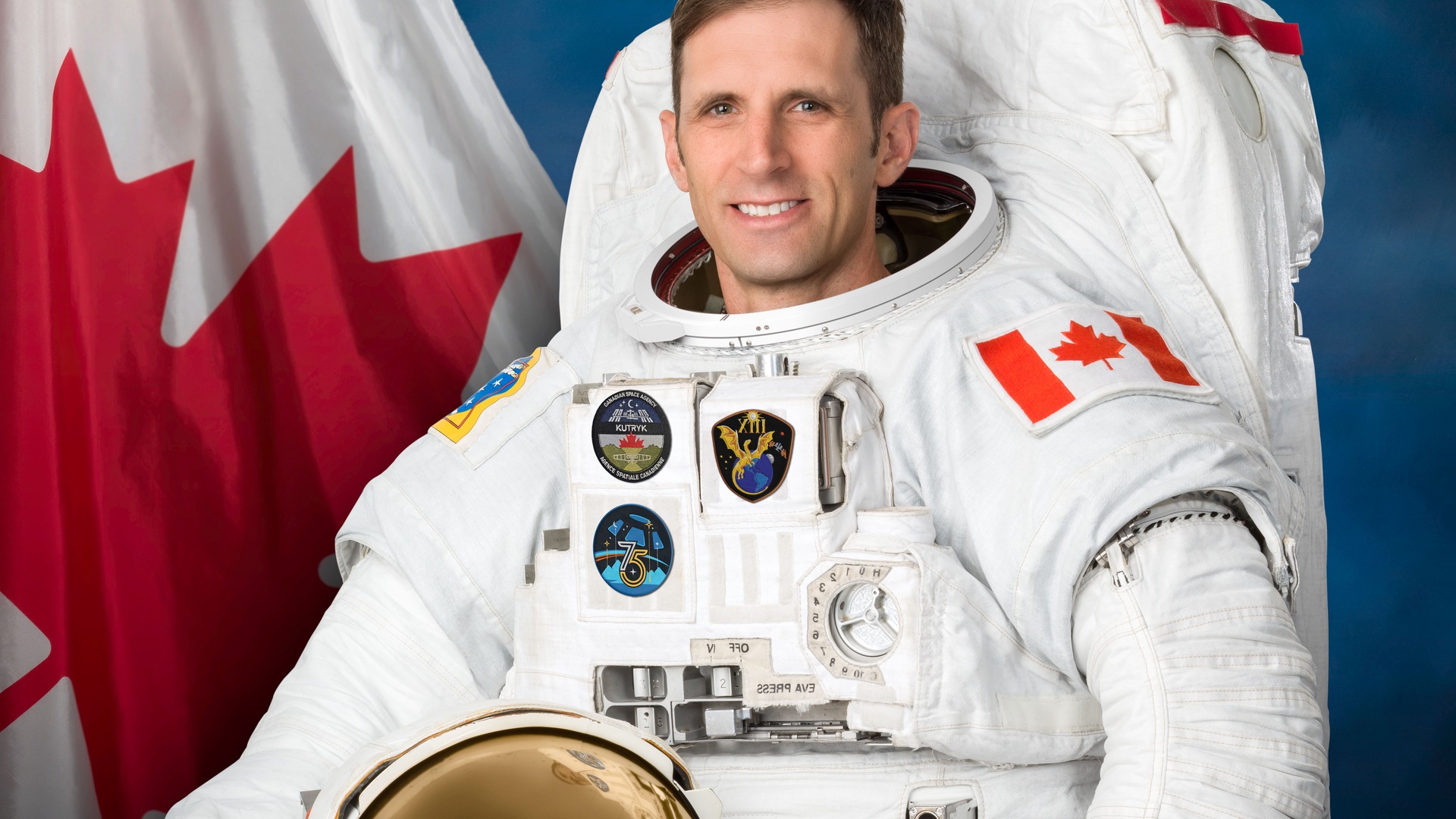 Canadian Space Agency astronaut Joshua Kutryk in a white spacesuit for his official Crew-13 portrait, holding his helmet with a Canadian flag behind him