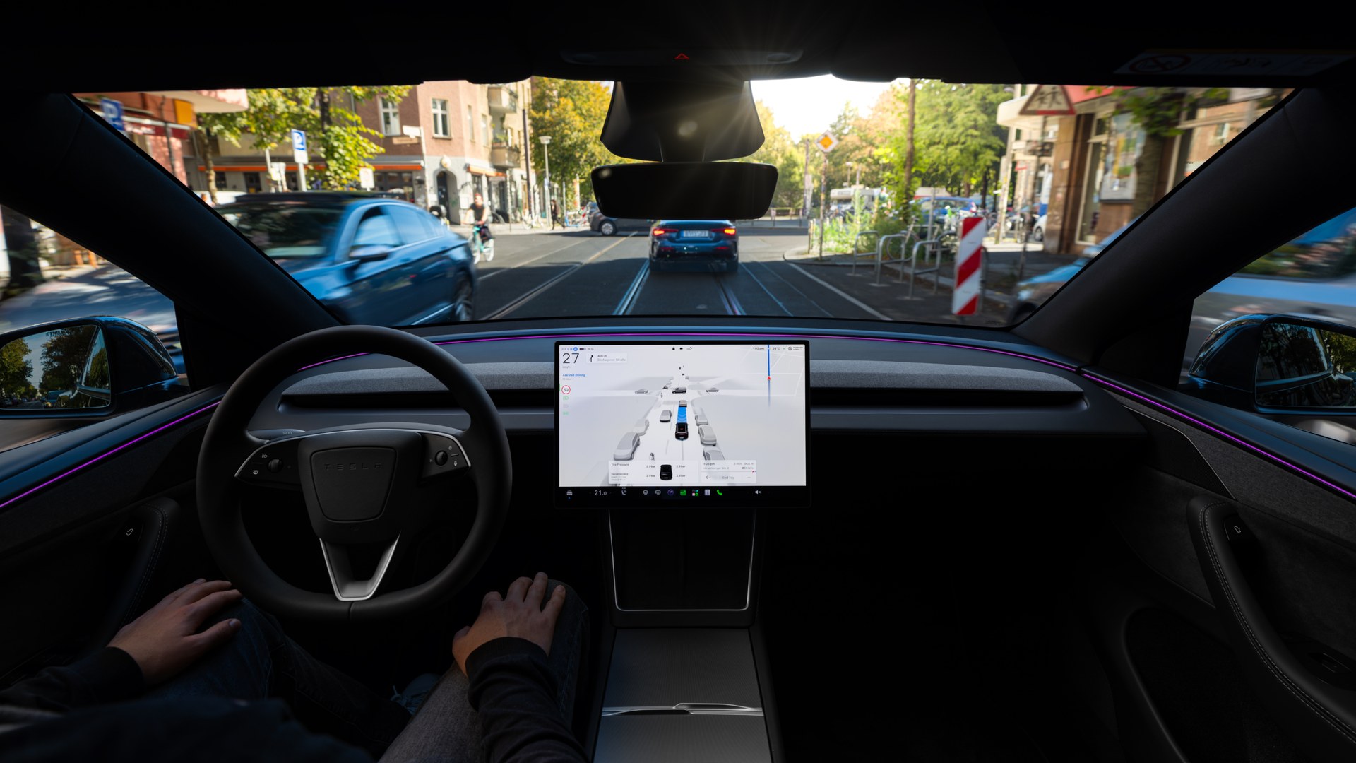 Interior view from the driver seat of a Tesla, with a large central touchscreen showing urban navigation ahead.
