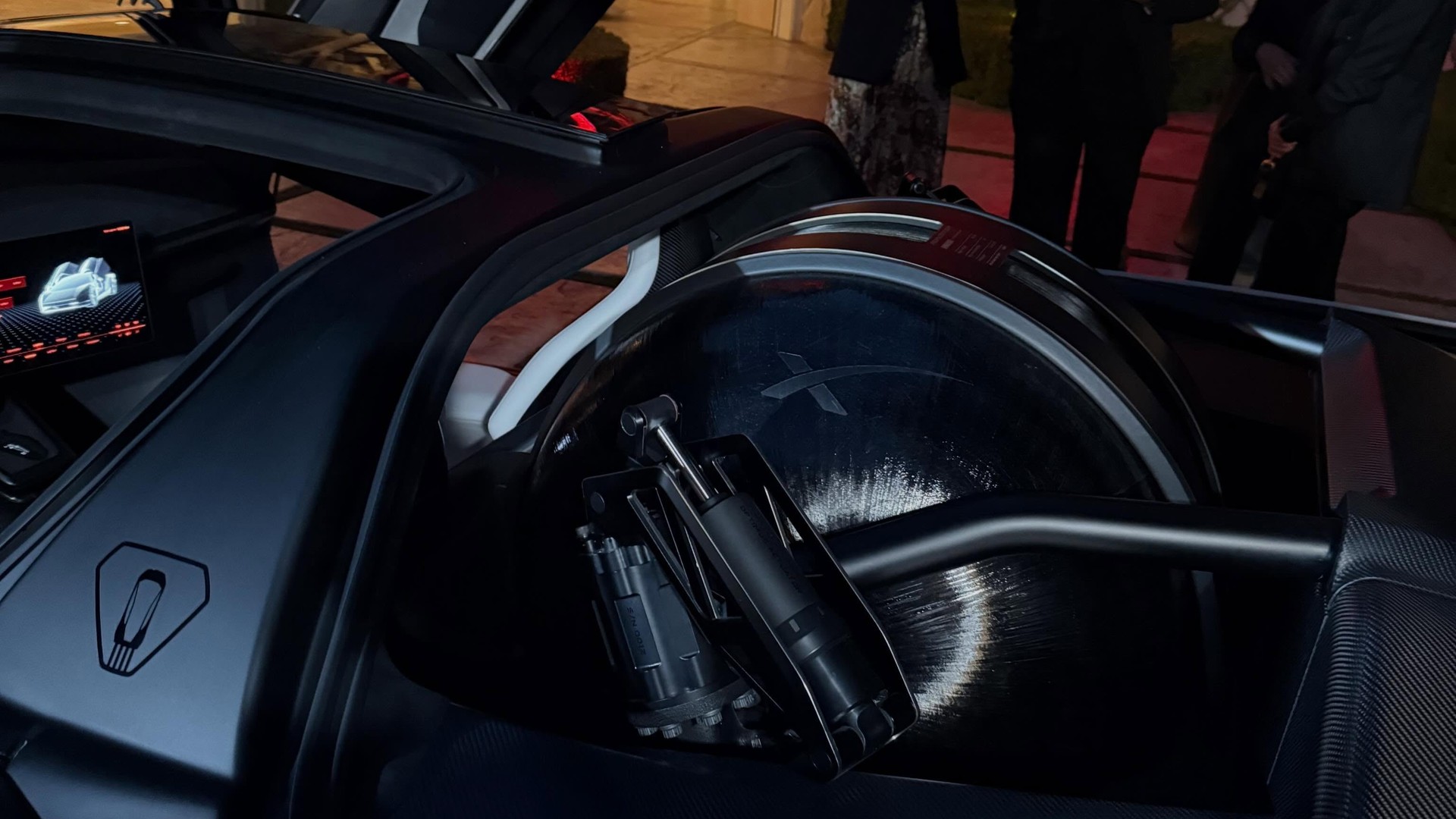 Close-up of a futuristic car interior showing a large circular mechanical module with a white support brace and a nearby touchscreen display; people stand in the background.