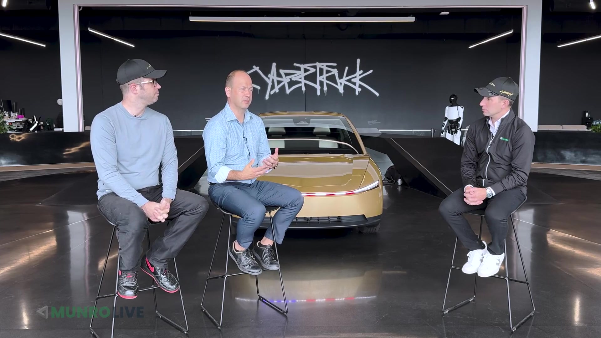 Lars Moravy speaks beside a gold Cybercab and Optimus robot in a Munro Live interview