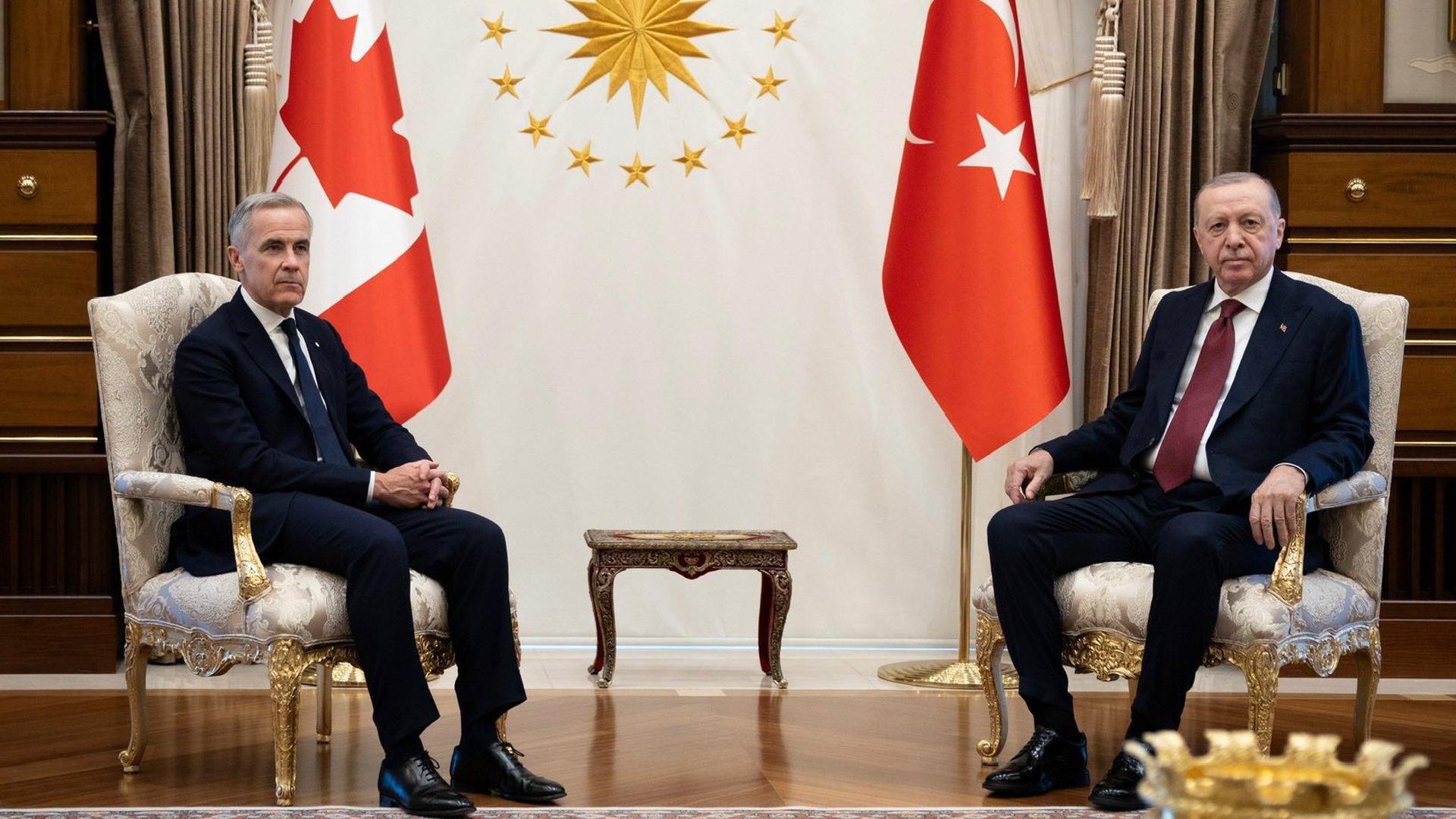 Two men in dark suits sit in ornate chairs in a formal meeting room, with Canadian and Turkish flags and a gold sun emblem behind them.