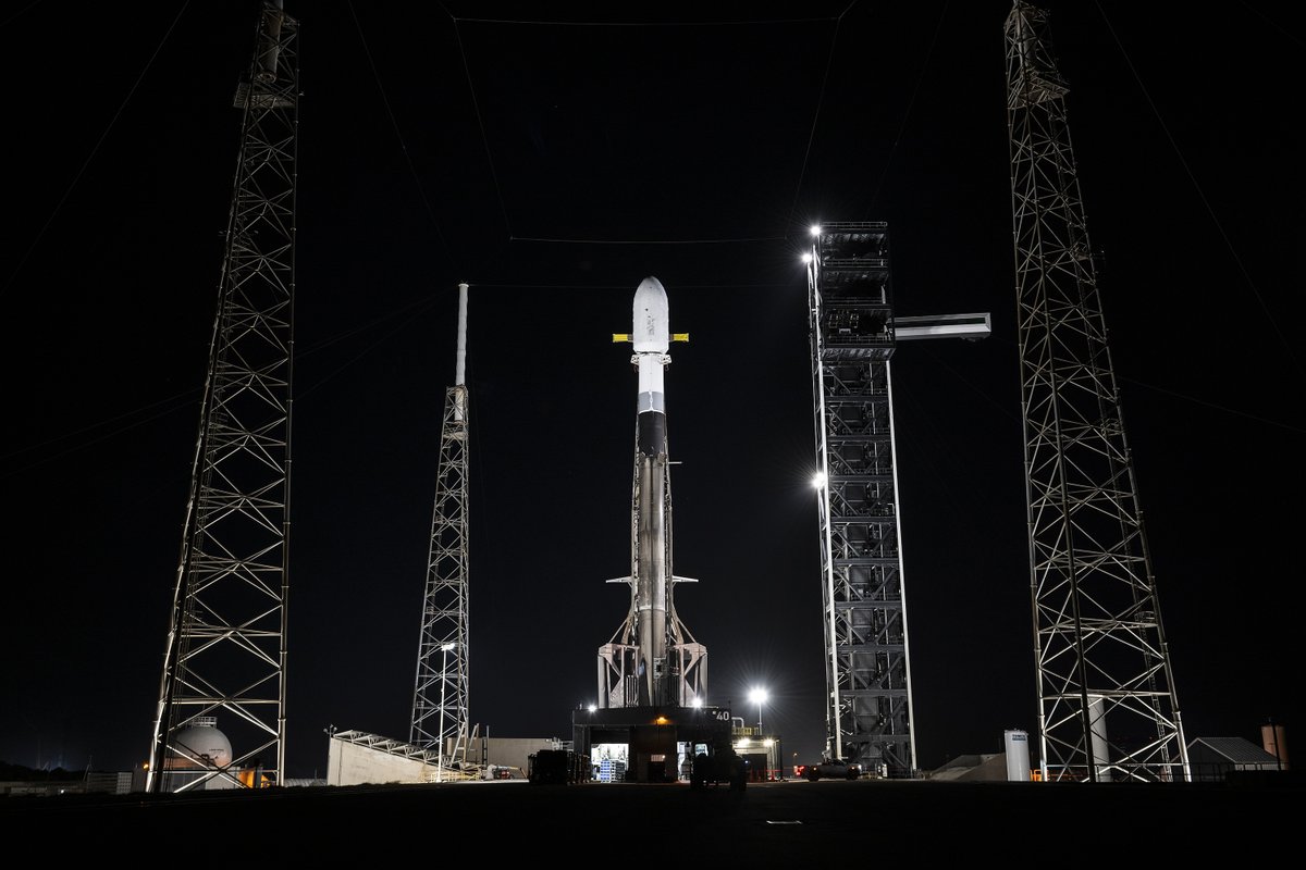 Rocket stands upright on a launch pad at night, framed by tall support towers and bright ground lights.