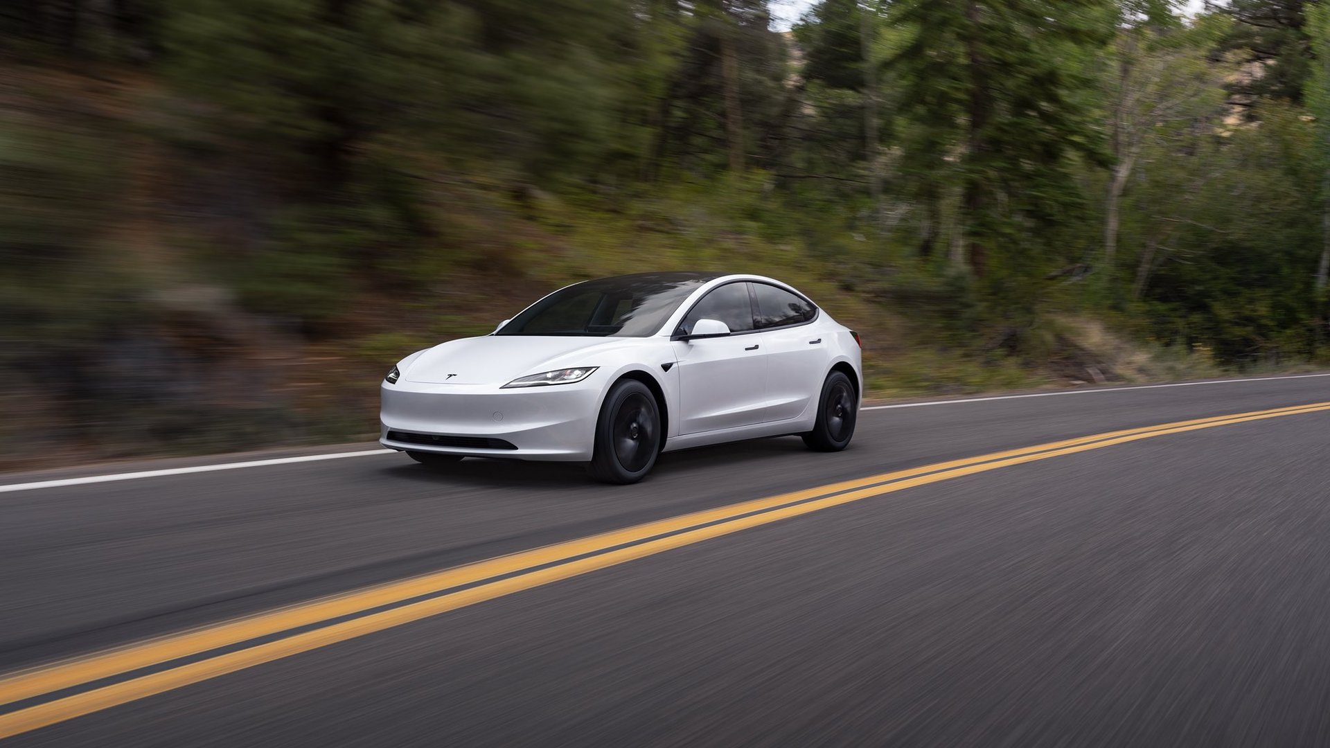 White Tesla Model 3 with black wheels driving on a two-lane road past a blurred forest