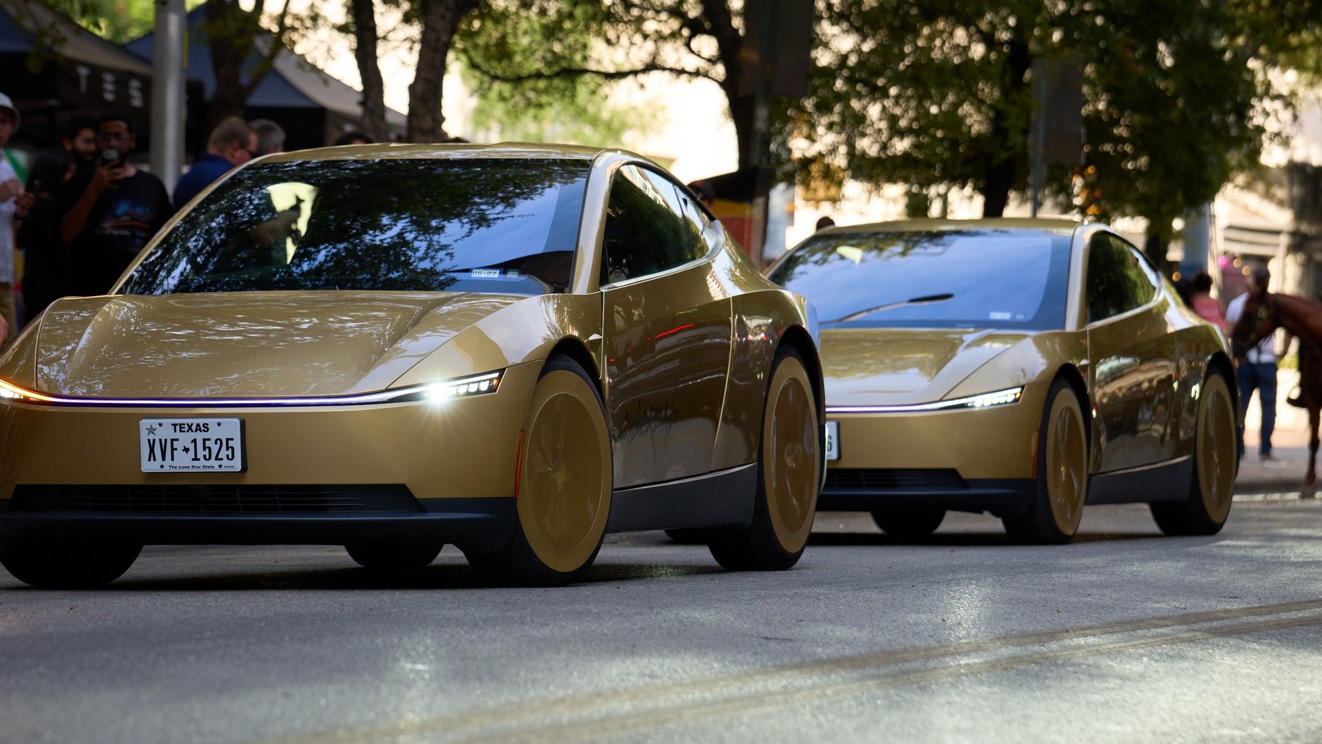 Two gold Tesla Cybercabs driving down a downtown Austin street past spectators and a mounted police officer