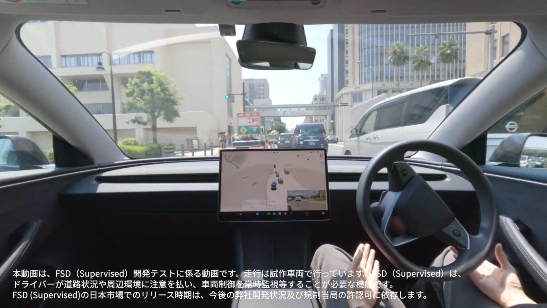 Front-seat car interior with a large central touchscreen showing navigation, hands on the steering wheel, city street visible through the windshield, and Japanese captions overlaying the video.