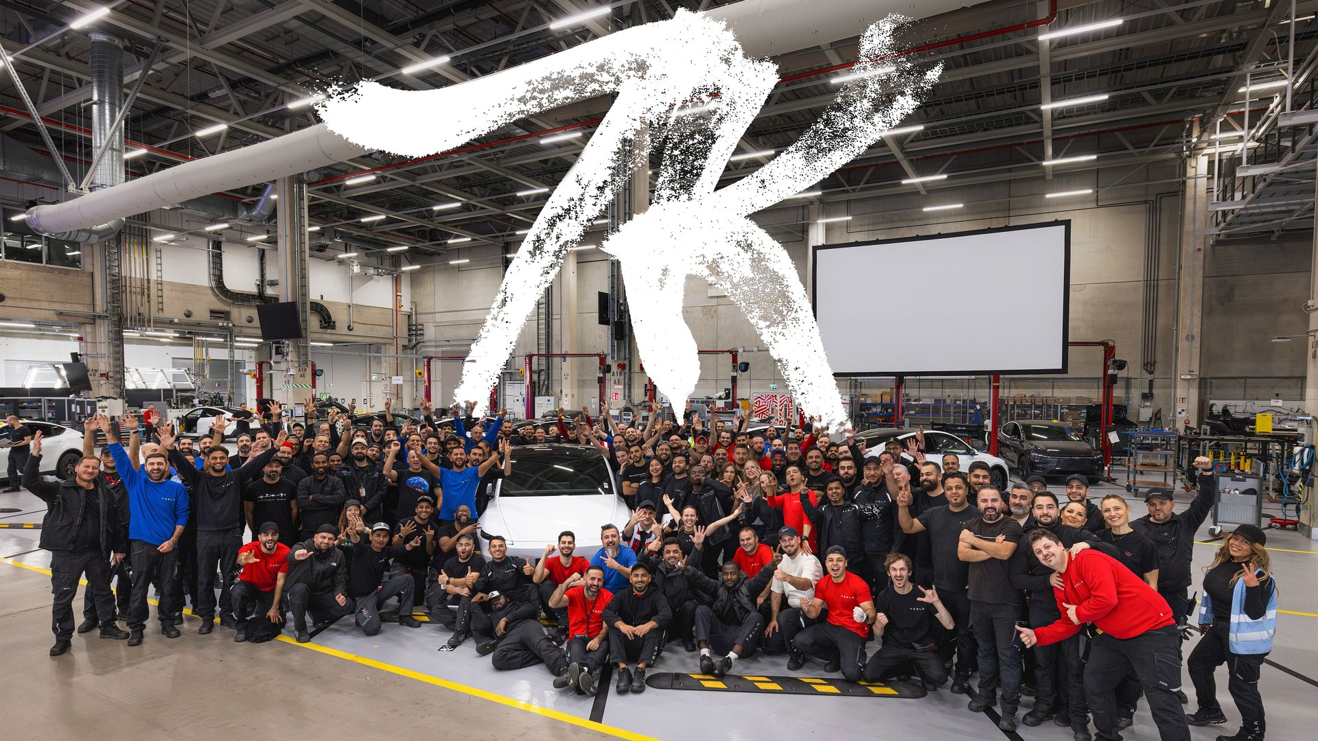 Giga Berlin workers celebrate around a white Tesla Model Y on the factory floor under a large painted 7K