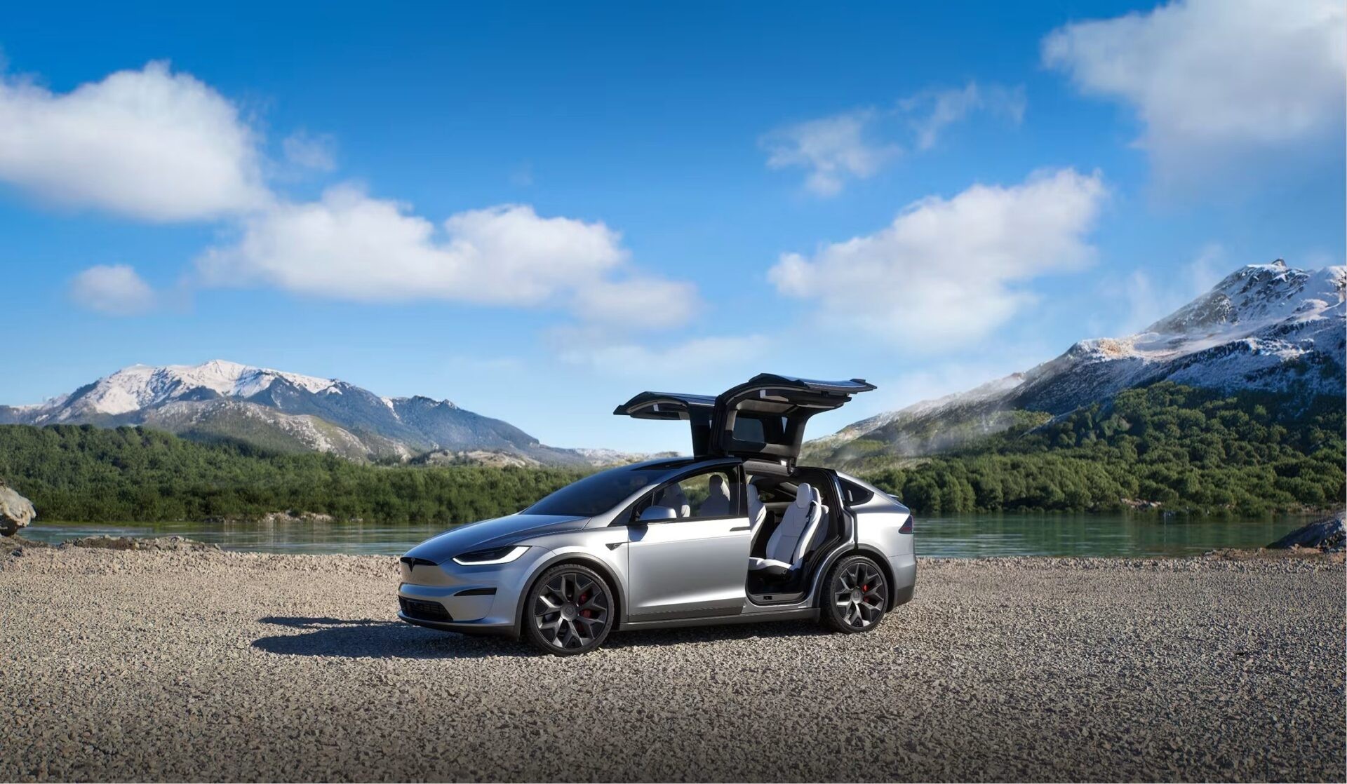 A silver Tesla Model X with its Falcon Wing doors open on a gravel shore beside a lake and snow-capped mountains