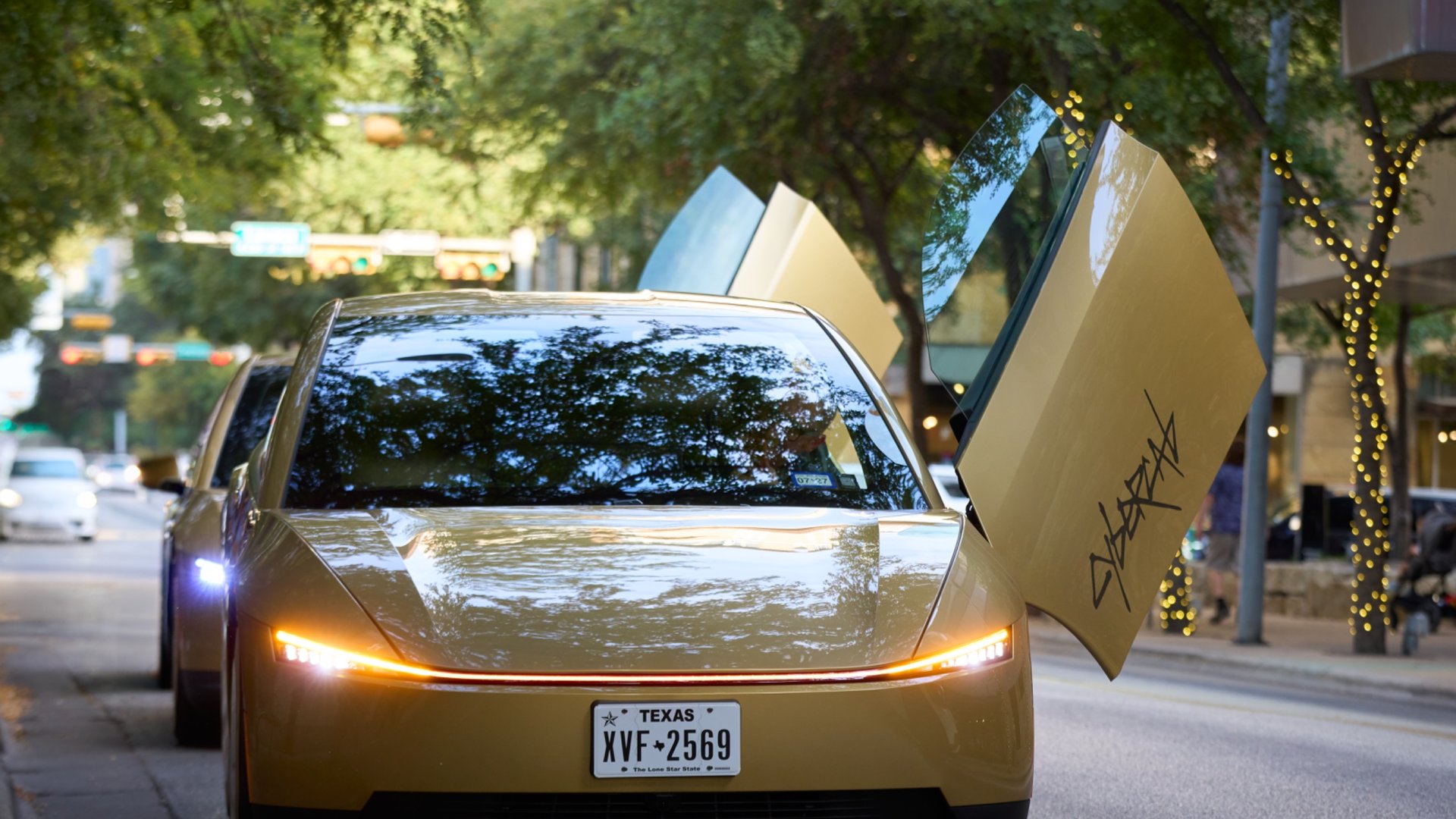 A gold Tesla Cybercab with its butterfly doors raised on a tree-lined downtown Austin street
