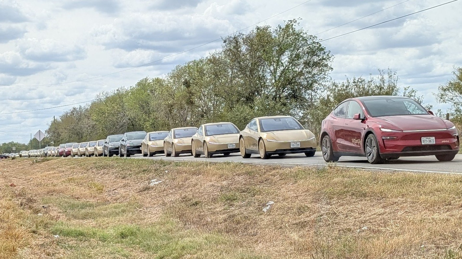 Line of many Tesla cars in different colors driving on a highway, with a red car at the front and a clear blue-sky backdrop.