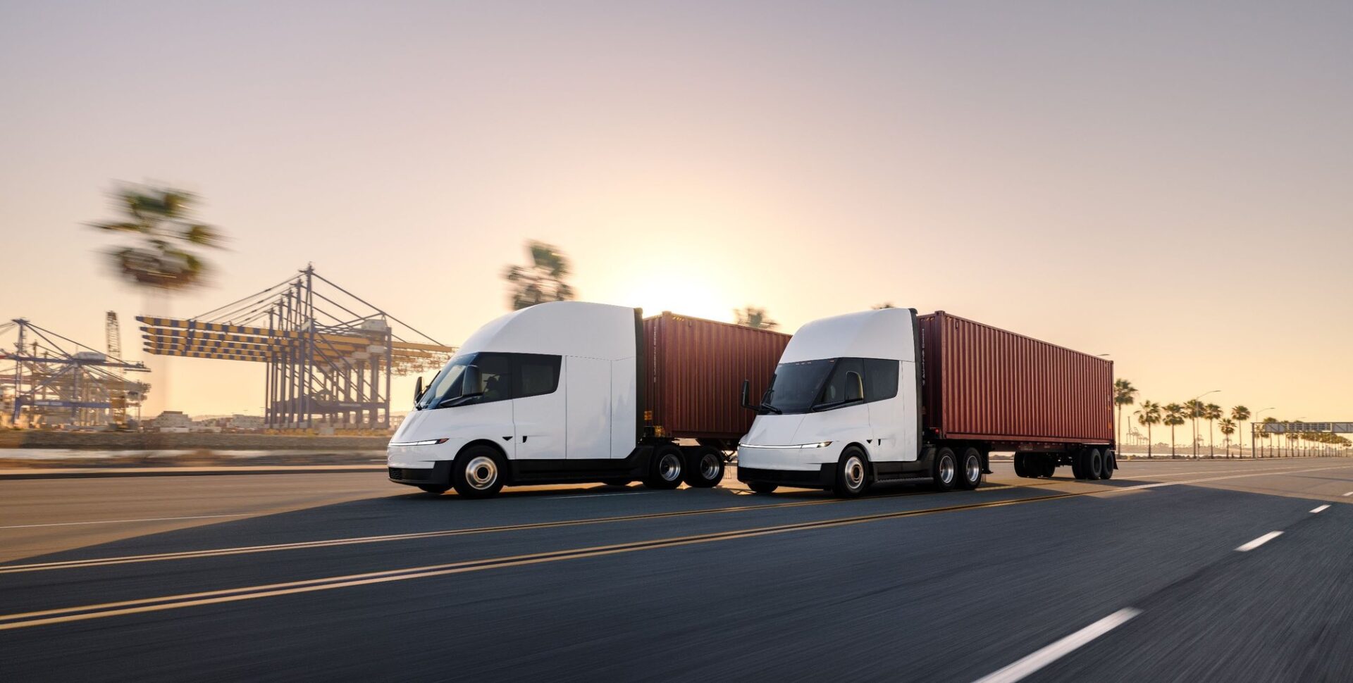 Two Tesla Semi trucks hauling shipping containers near a port at sunset