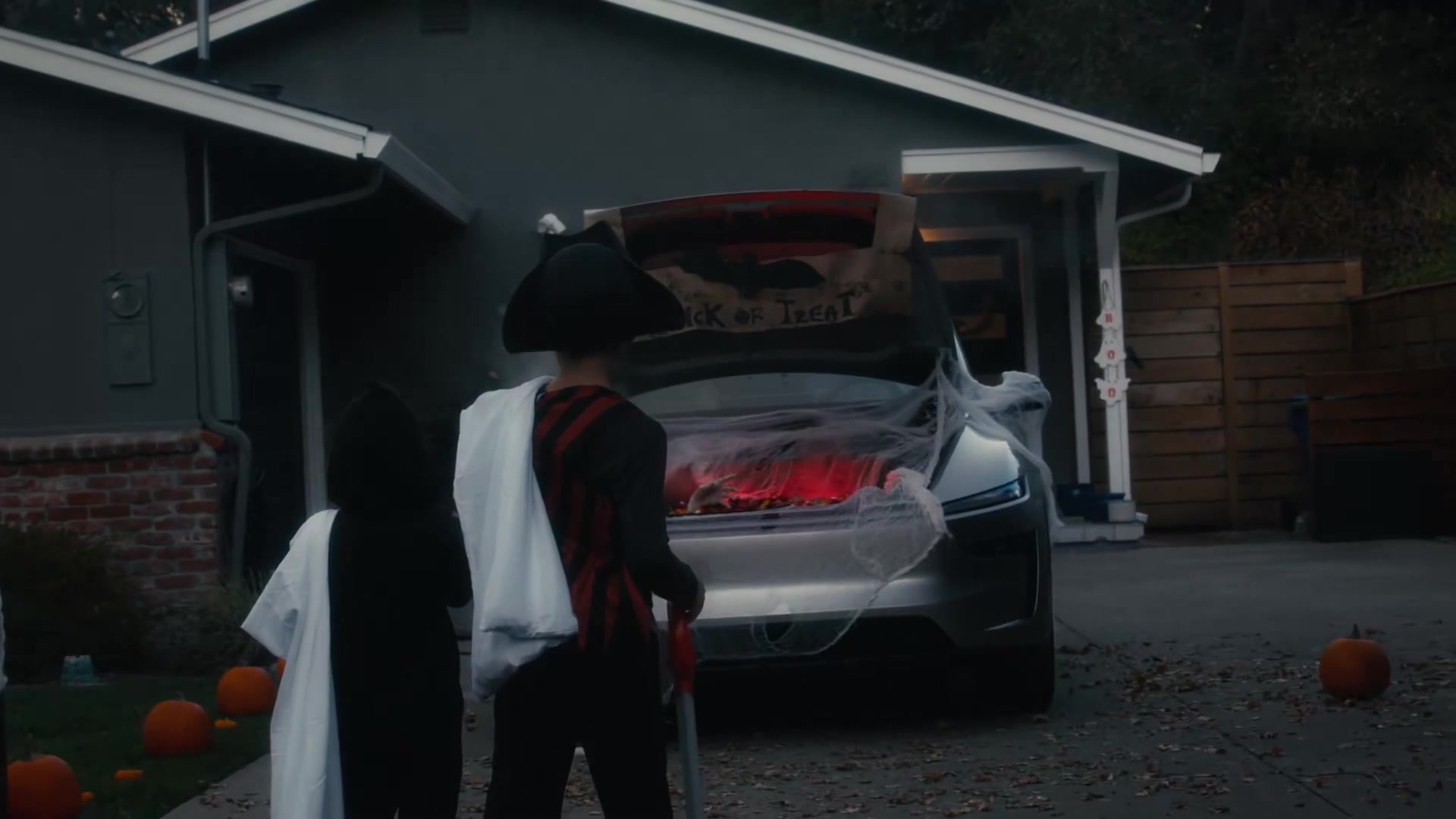 Silver Tesla with red-lit open trunk and Trick or Treat sign as costumed kids approach at dusk
