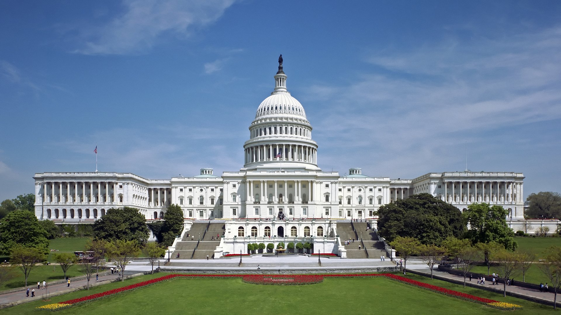 The U.S. Capitol building with a domed roof, white stone, and surrounding lawn under a clear blue sky.